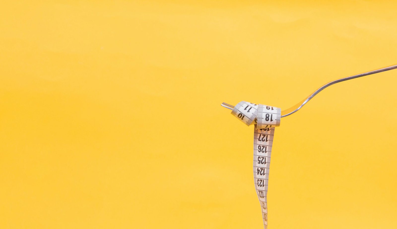 A photo of a fork with a measuring tape wrapped around it like spaghetti, against a bright yellow background. (How many calories are in a measuring tape?)