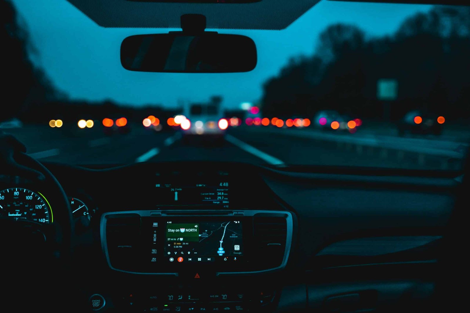 Close up view of a car dashboard showing the speedometer and a GPS system showing a route on the New Jersey Turnpike, starting place for many road trips. A highway is visible in the background, through the car's windshield. It's dusky outside.