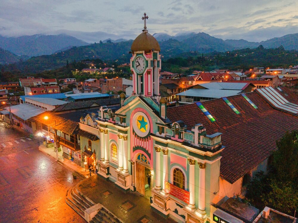 The featured images for "Las Cruces: Flash Fiction": A large church is painted bright pink and green and is lit up with multicolored lights. It faces a cobblestone plaza; the colorful lights are reflected on the wet cobblestones. Tree-covered mountains are in the background.