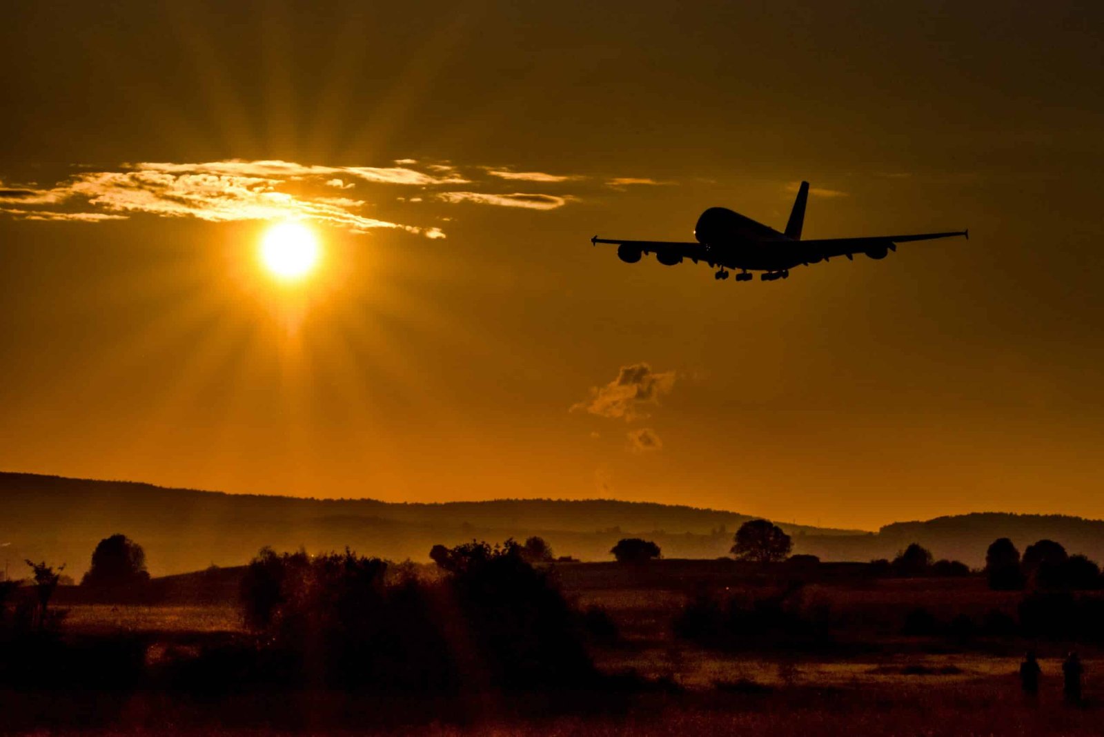 An airplane is in silhouette against the setting sun as it flies over a city.